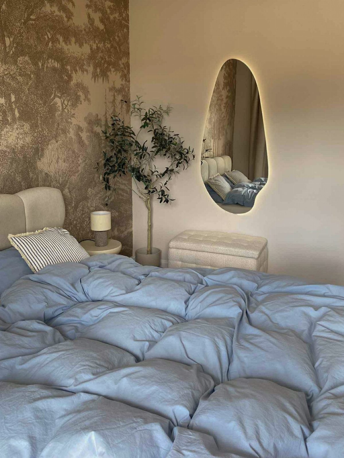 Bed with blue comforter next to a tree-patterned wall and oval mirror.