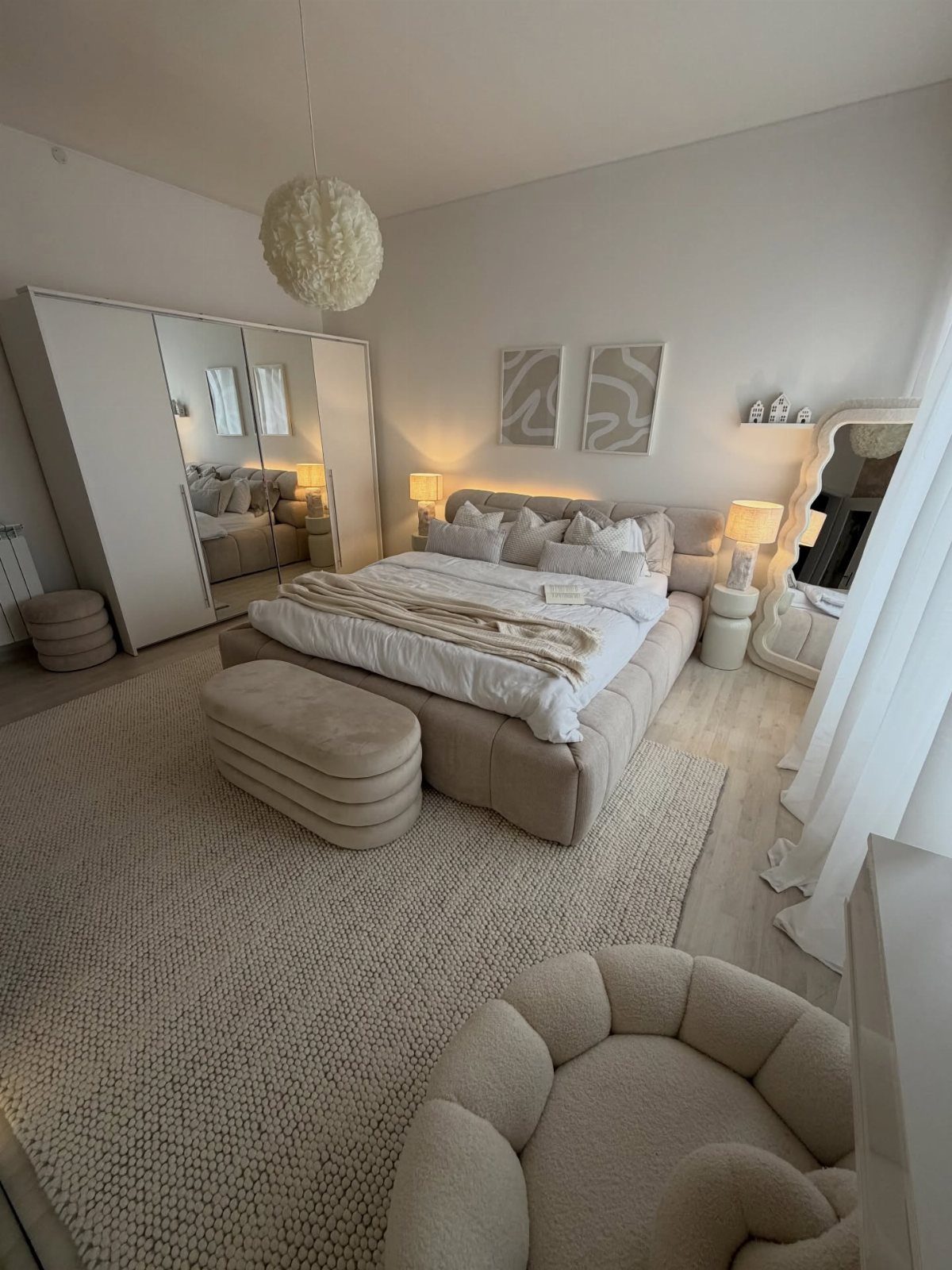 Bright monochromatic bedroom with cream upholstered bed, textured rug, and fluffy pendant light.