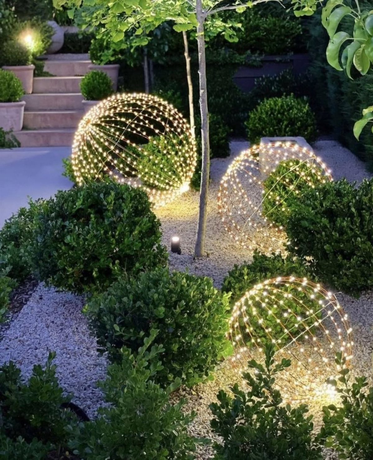 Glowing spherical LED light installations placed among green shrubs in a gravel garden.