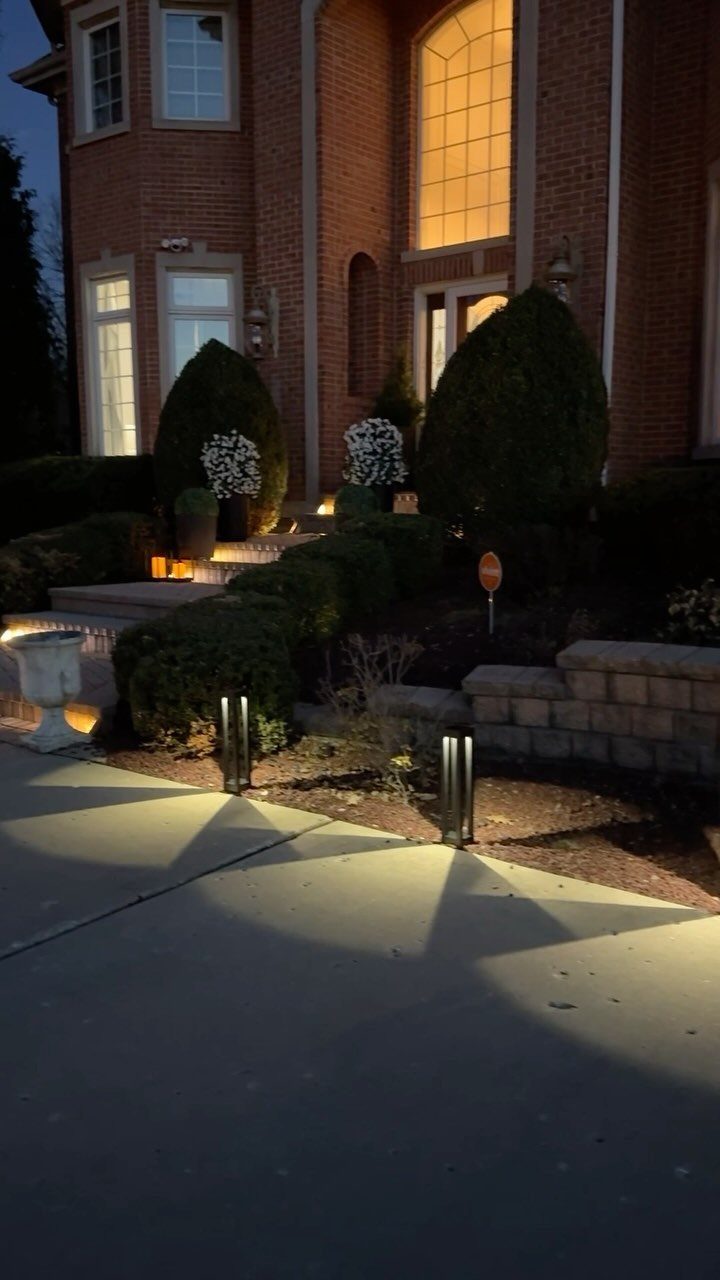 Black cylindrical path lights illuminating a concrete walkway in front of a brick house