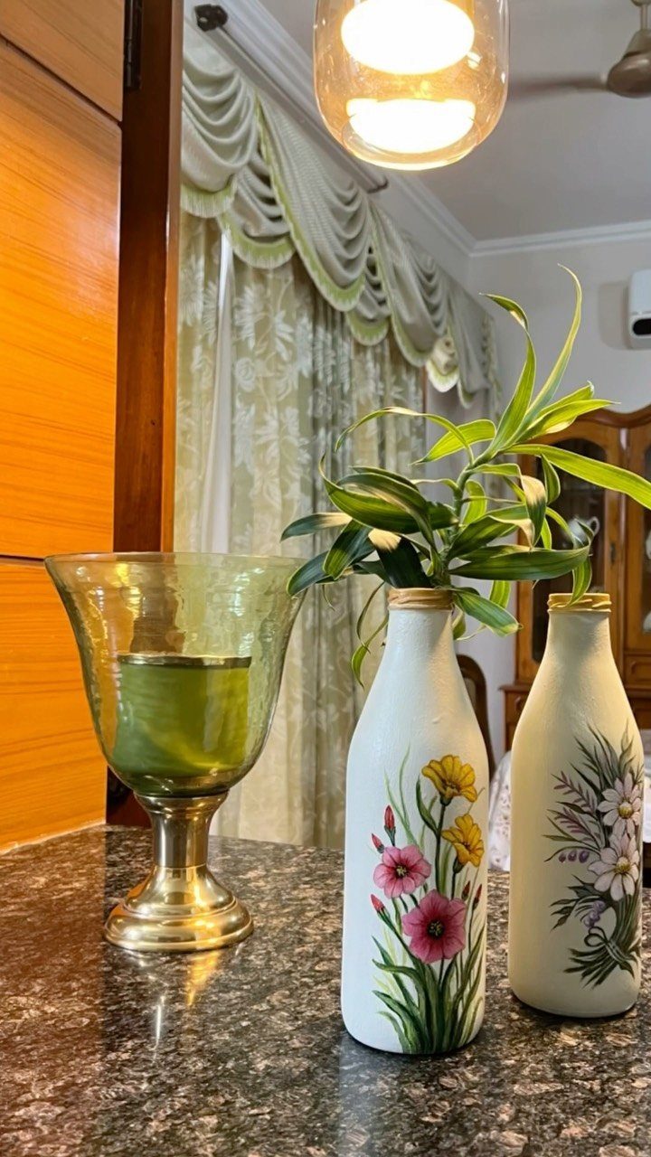 Two white painted bottles with floral art and a green glass goblet on granite.