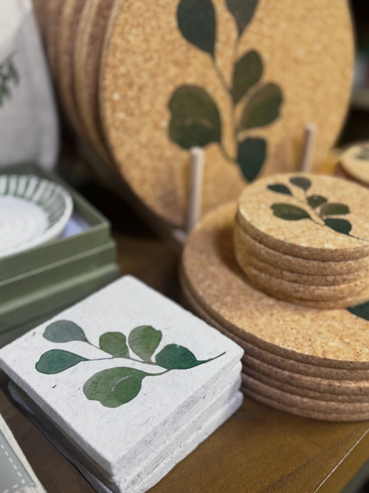 Circular cork and square stone coasters with green leaf prints on a wooden table
