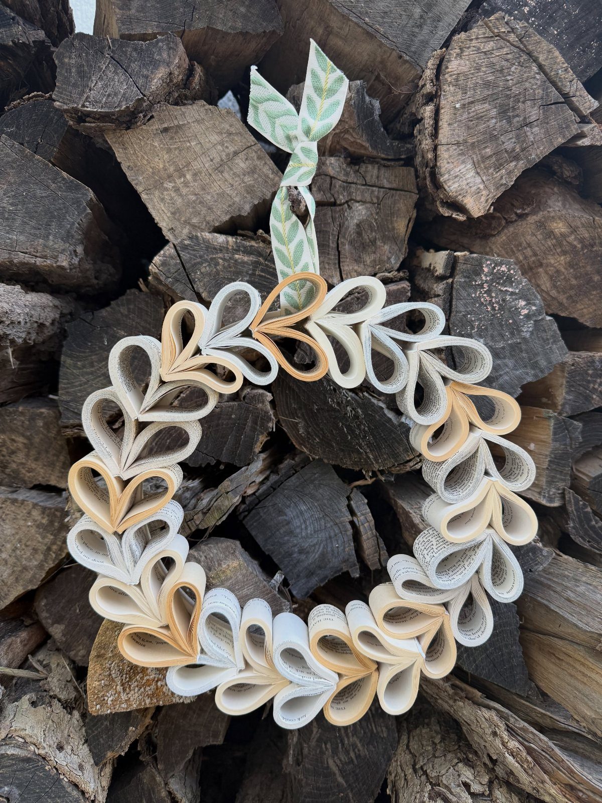 Paper wreath made from folded book pages hanging against a stack of firewood.