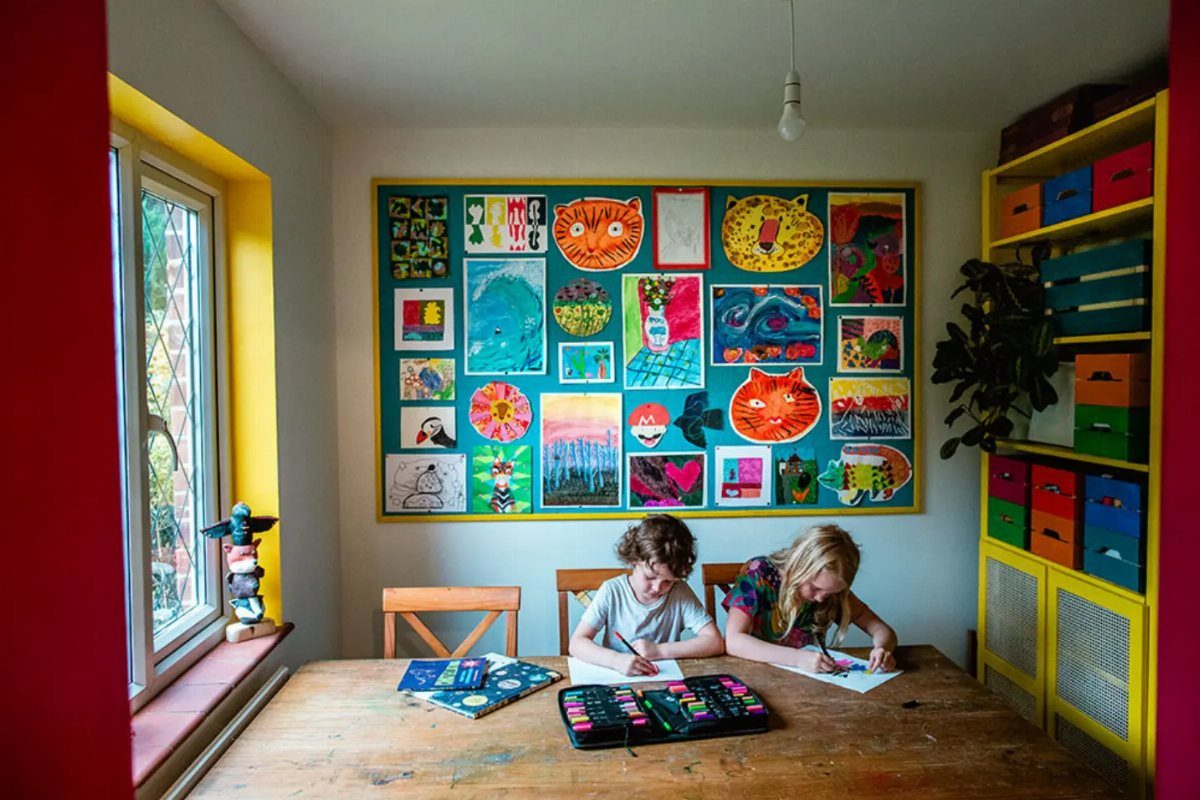 Children drawing at a table before a large yellow-framed corkboard with colorful art.