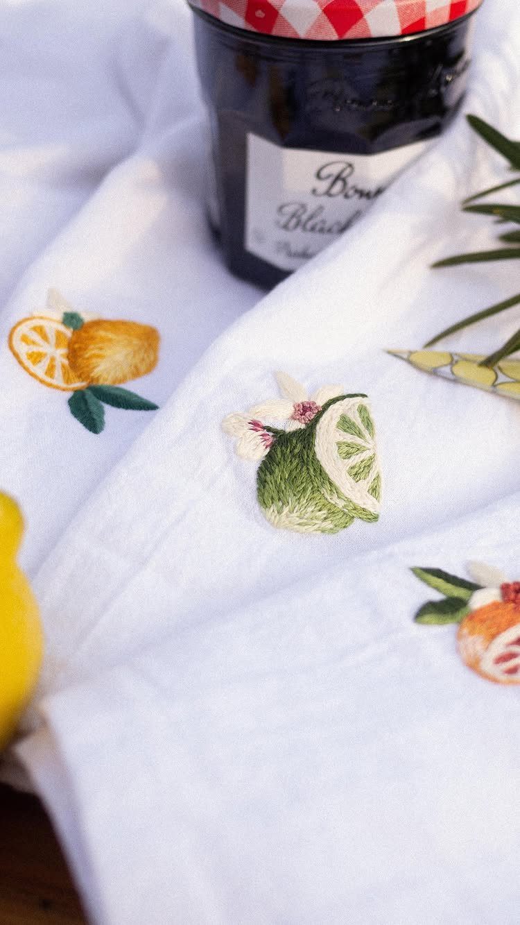 White kitchen towel with embroidered citrus fruits next to a dark glass jar.
