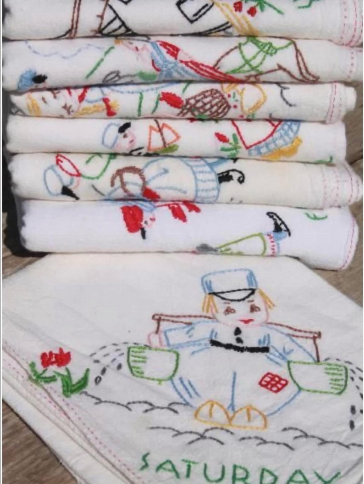 Stacked white flour sack towels with colorful embroidered characters and the word Saturday.