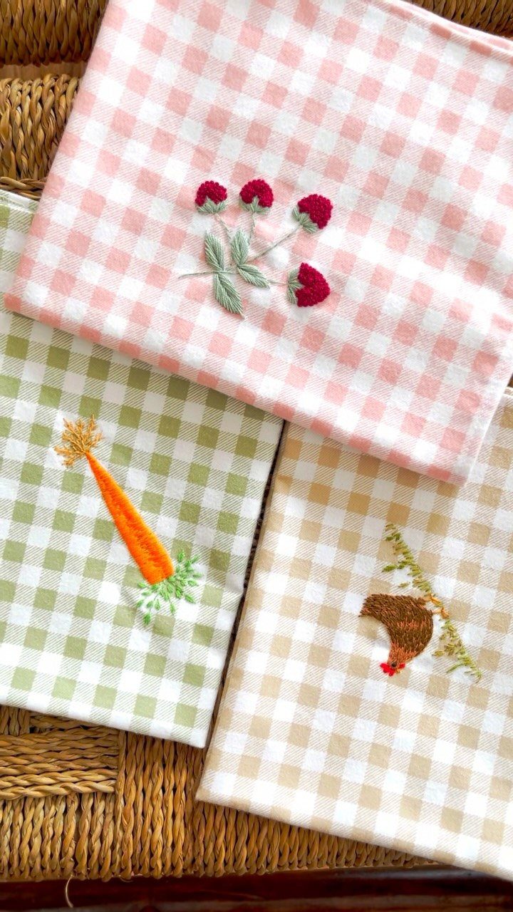 Checkered kitchen towels with embroidered berries, a carrot, and a mushroom on wicker.