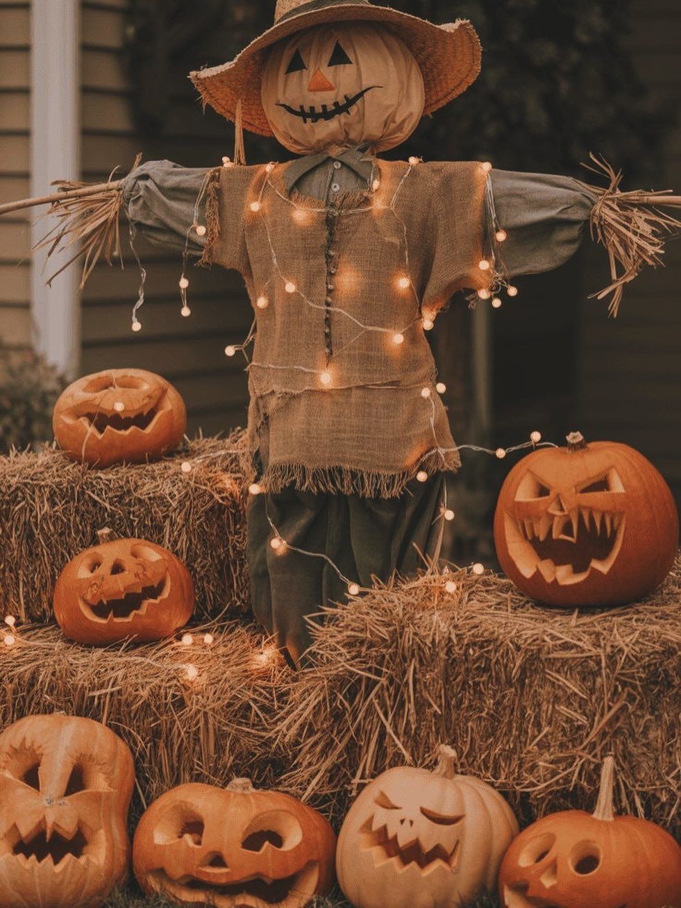 Scarecrow with pumpkin head and straw hat standing among hay bales and jack-o'-lanterns.