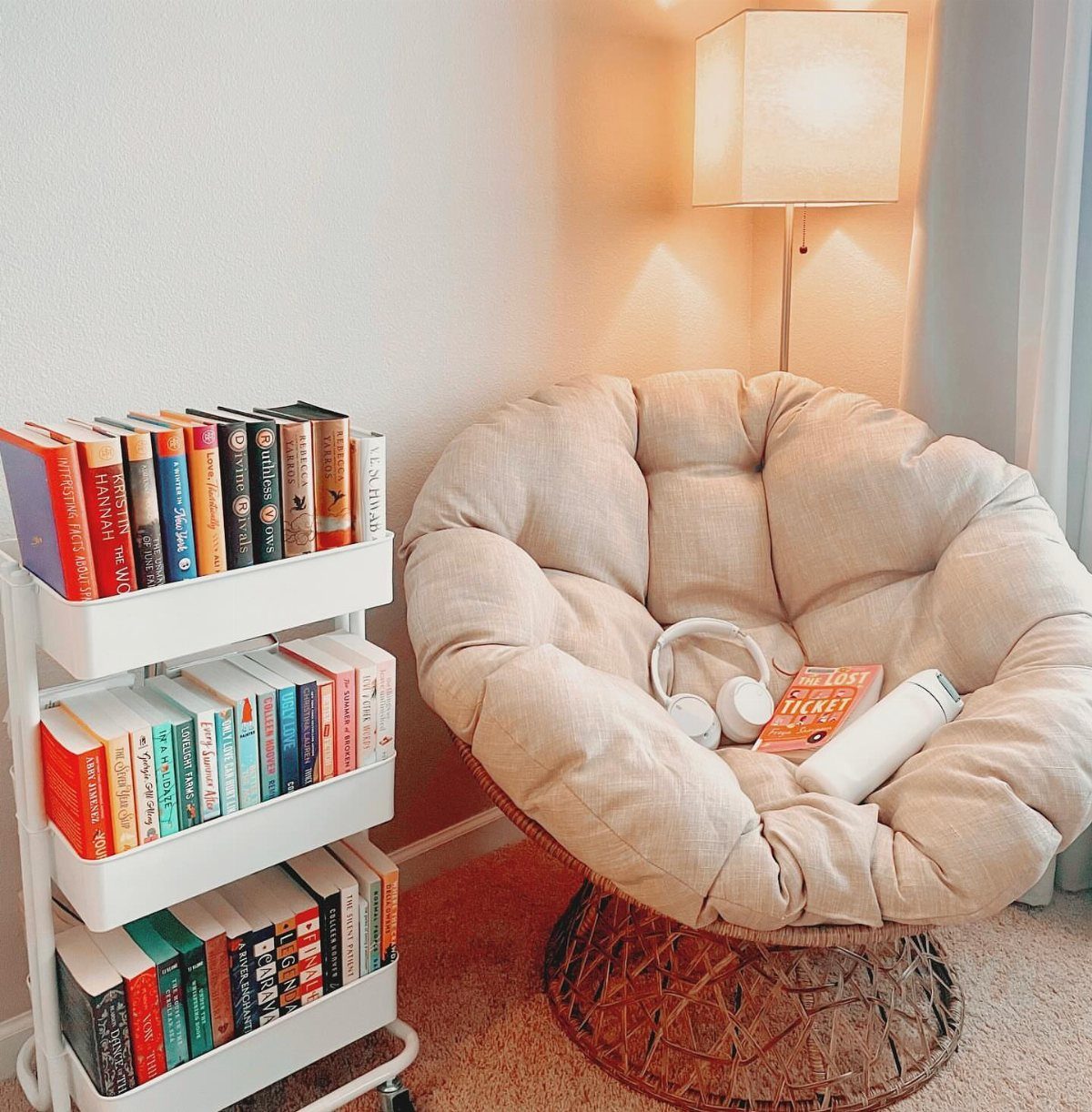 Beige papasan chair with a white rolling book cart and a lit floor lamp.