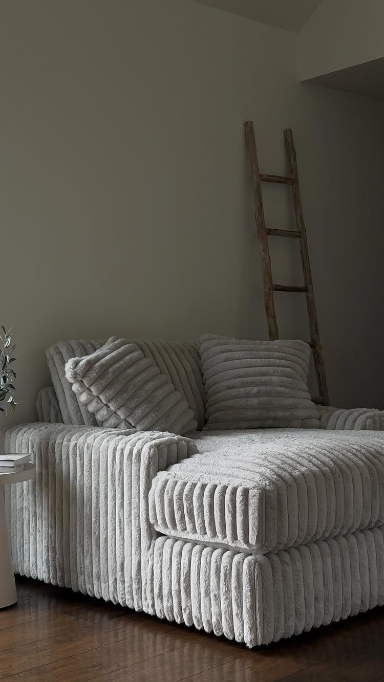 Grey ribbed fabric chaise lounge with pillows against a beige wall and wooden ladder.