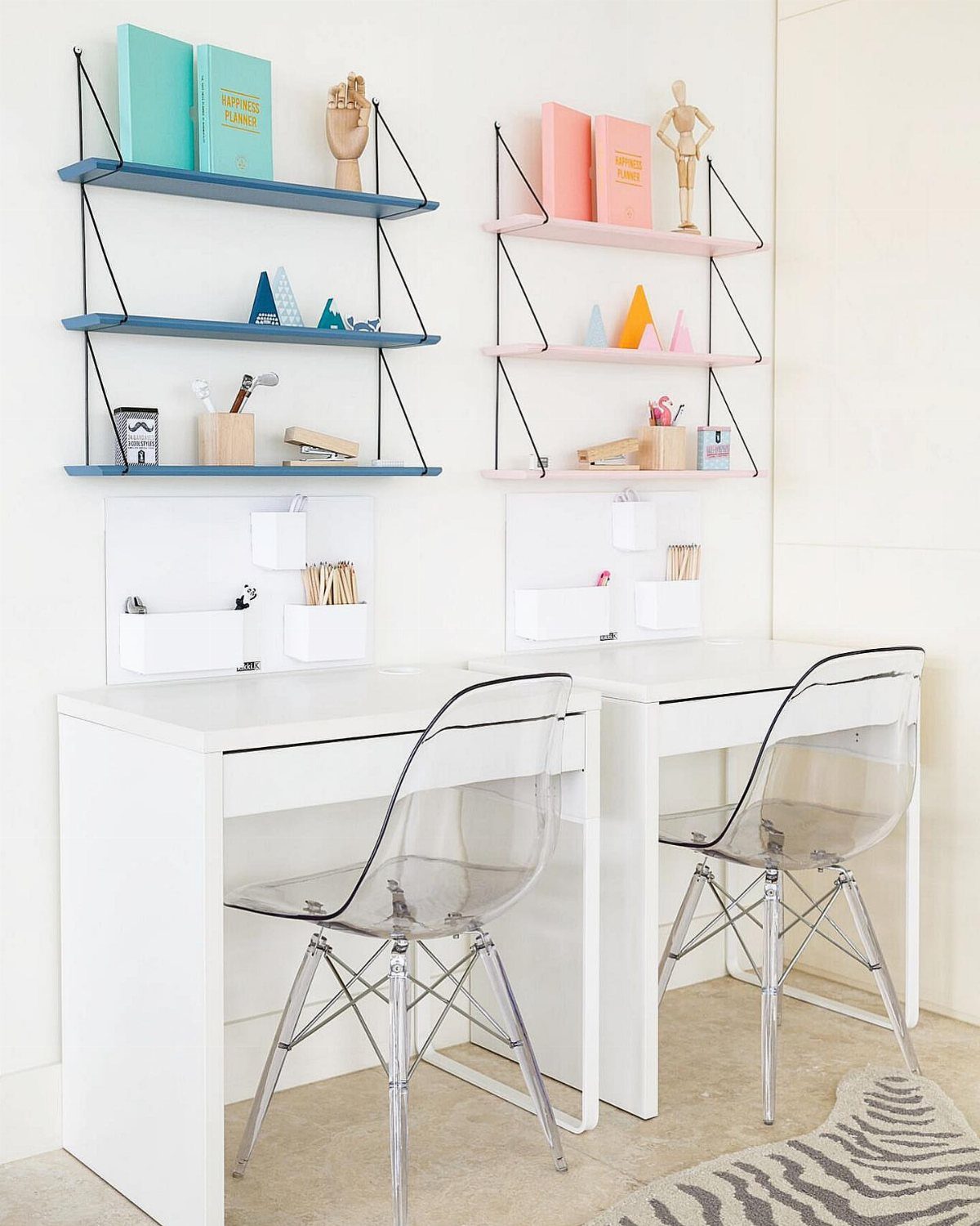 Two white desks with clear acrylic chairs and colorful floating wall shelves above them.