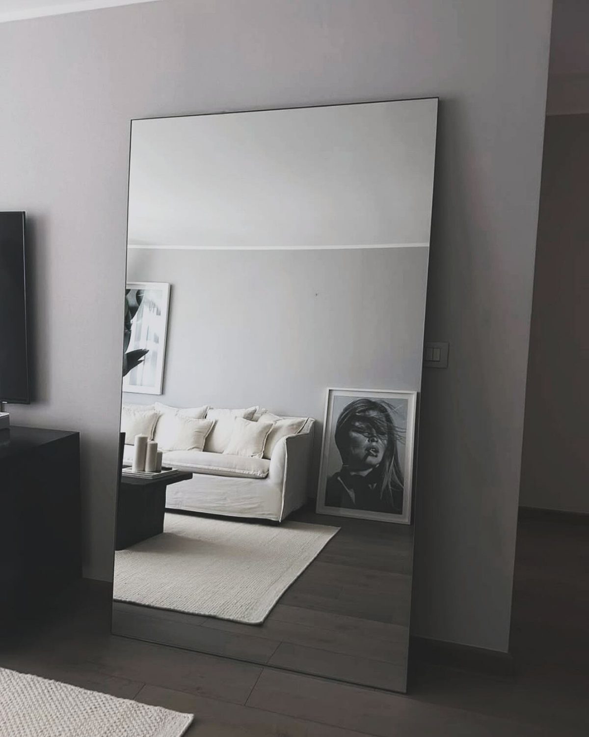 Large frameless floor mirror leaning against a wall reflecting a modern white living room