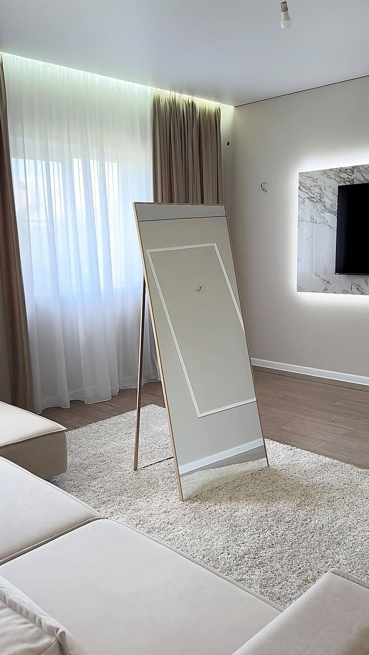 Large gold-framed floor mirror leaning in a bright living room with white rug.