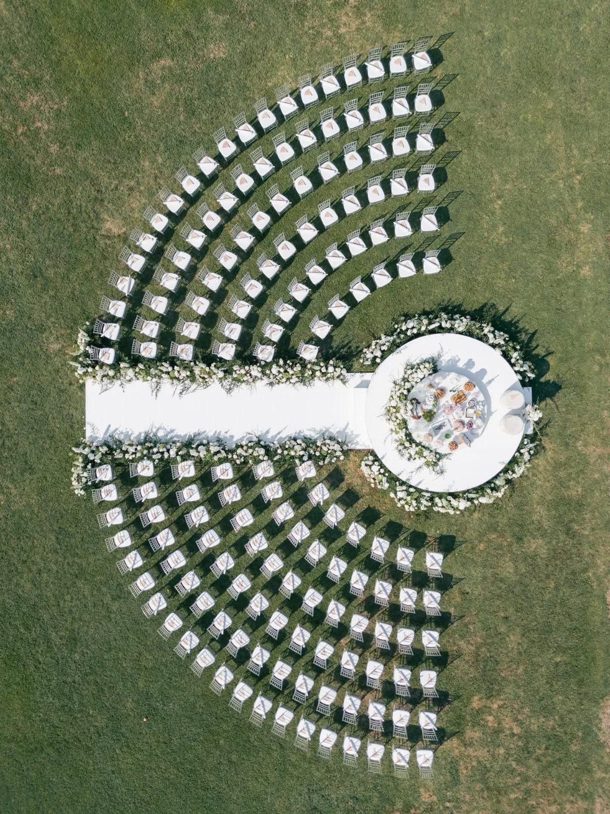 Curved rows of white chairs facing a circular floral wedding altar on grass.