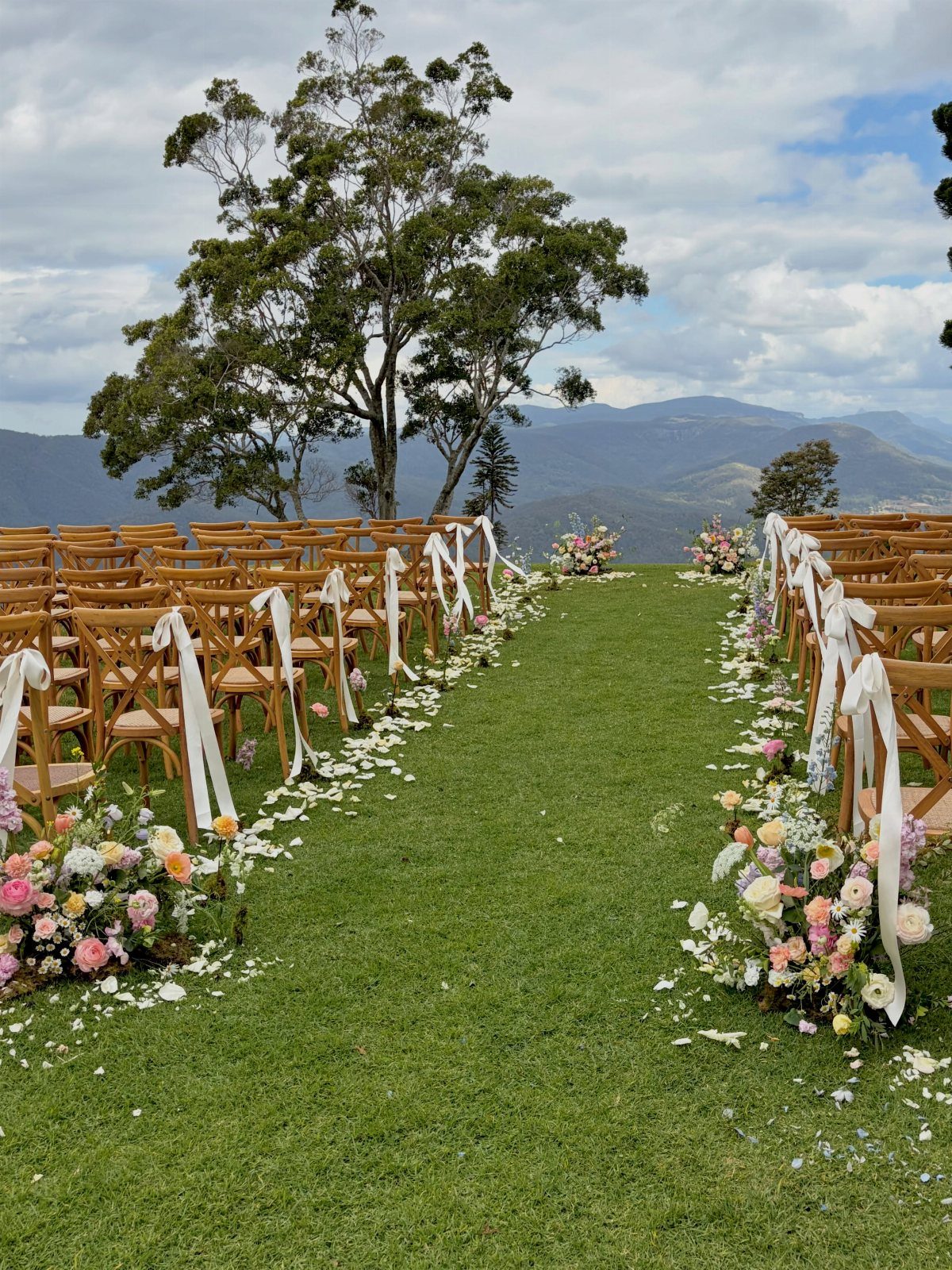Wooden wedding chairs with white ribbons lining a grassy aisle with floral arrangements.