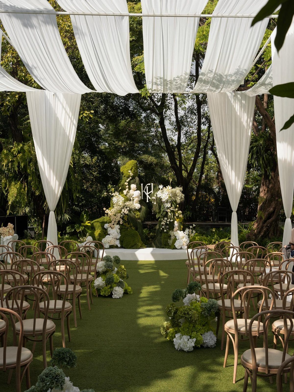 Outdoor wedding ceremony setup with white fabric draping, bentwood chairs, and white floral arrangements.