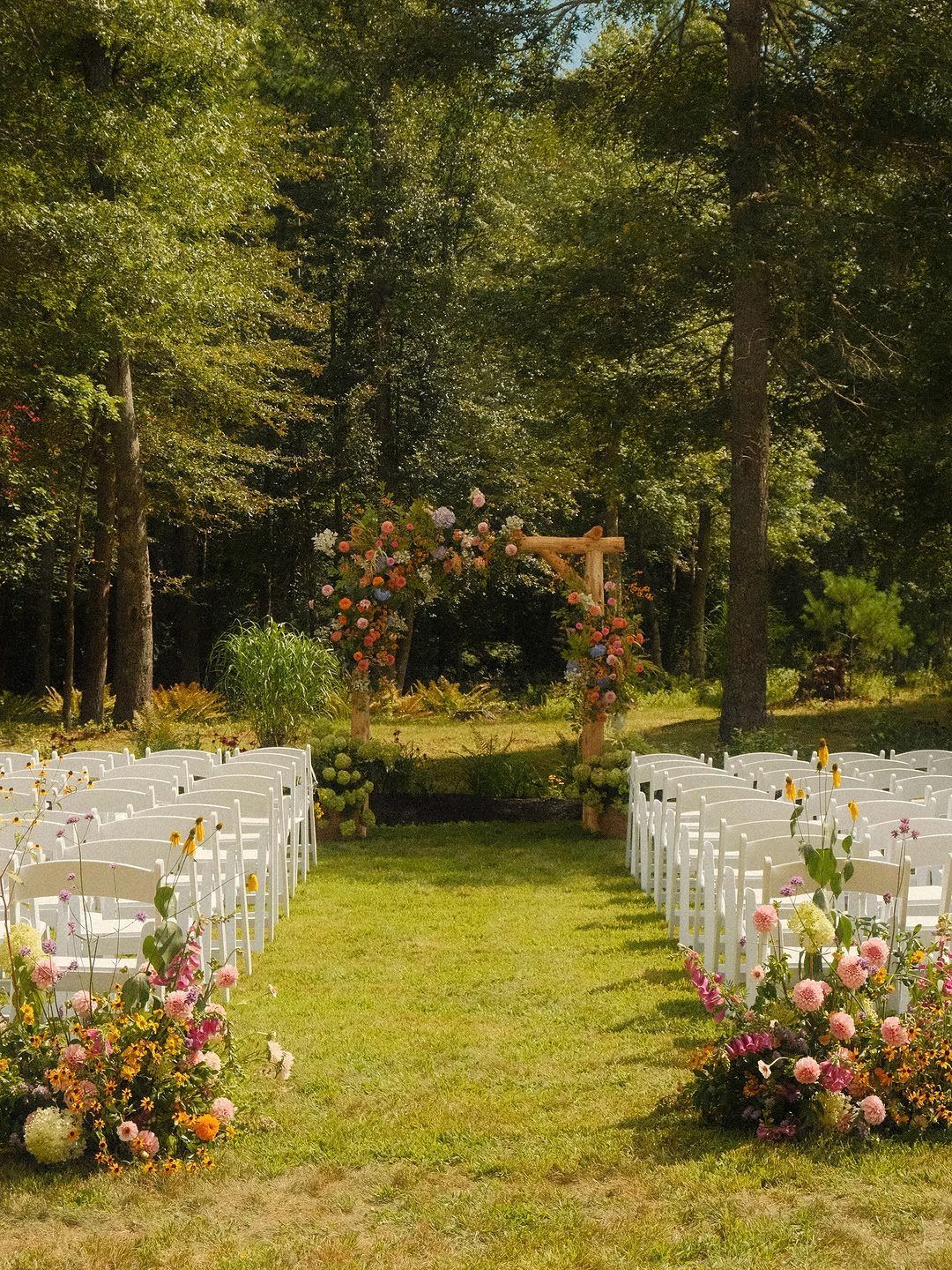 Outdoor wedding ceremony setup with white chairs and a floral wooden arbour in woods