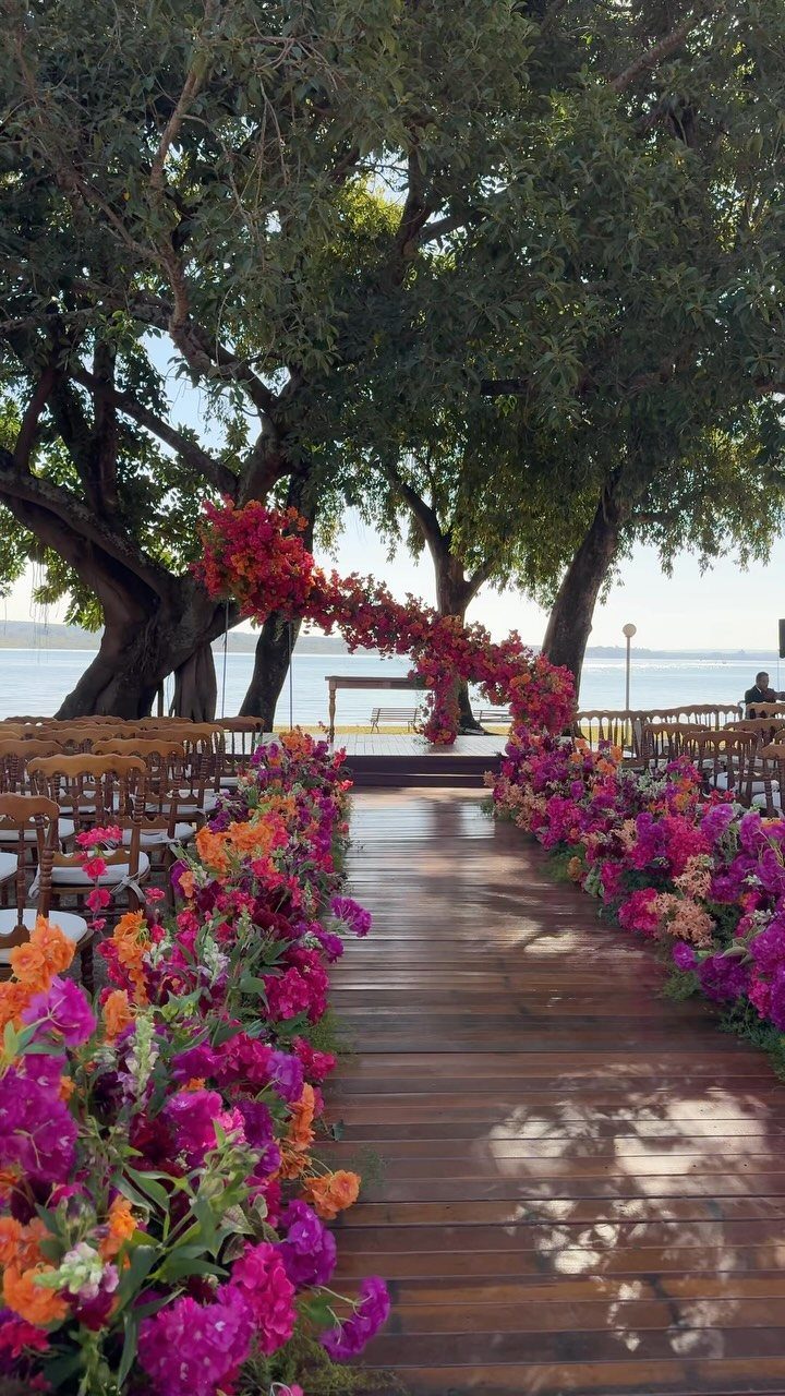 Wooden wedding aisle lined with colorful flowers under large trees by the water.