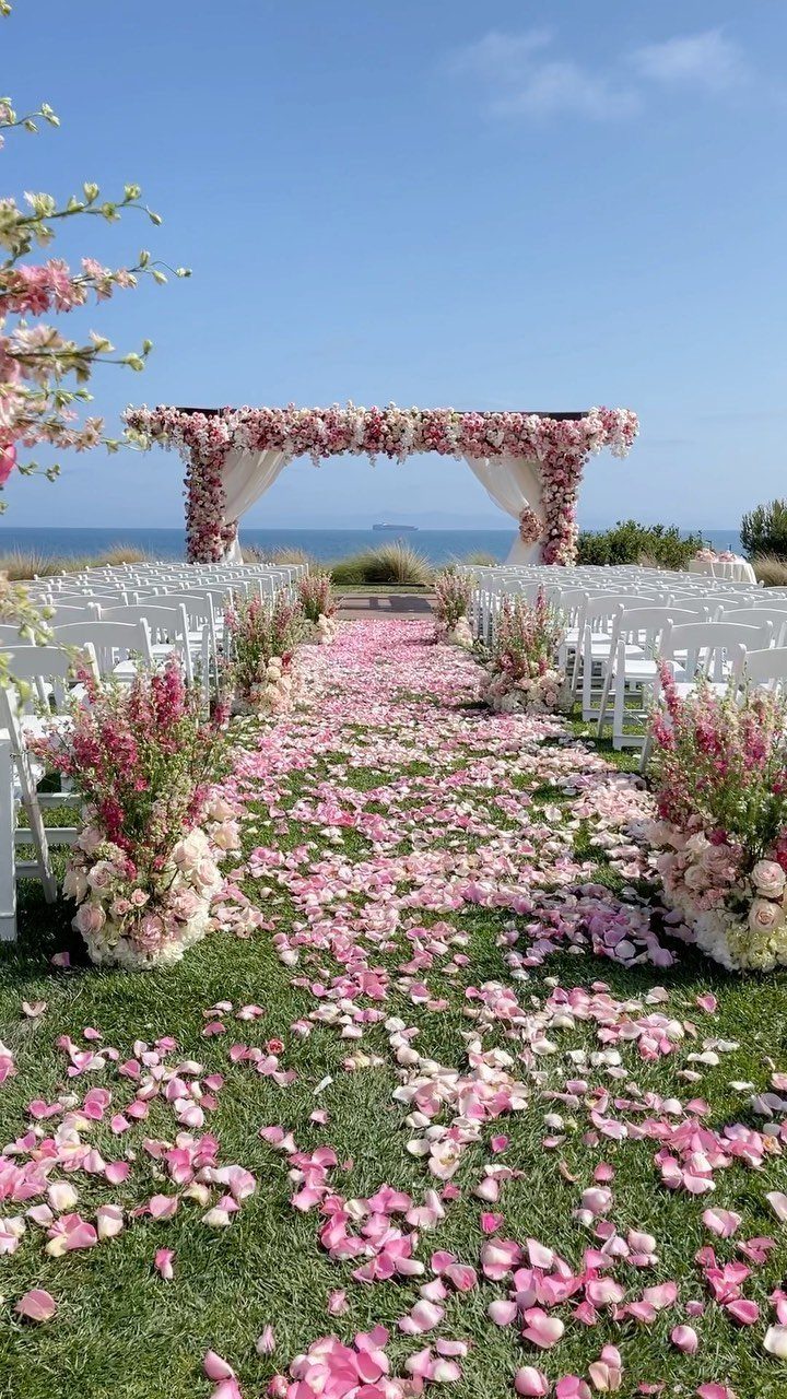 Pink flower-covered wedding arbour and petal-strewn aisle overlooking a blue ocean horizon.
