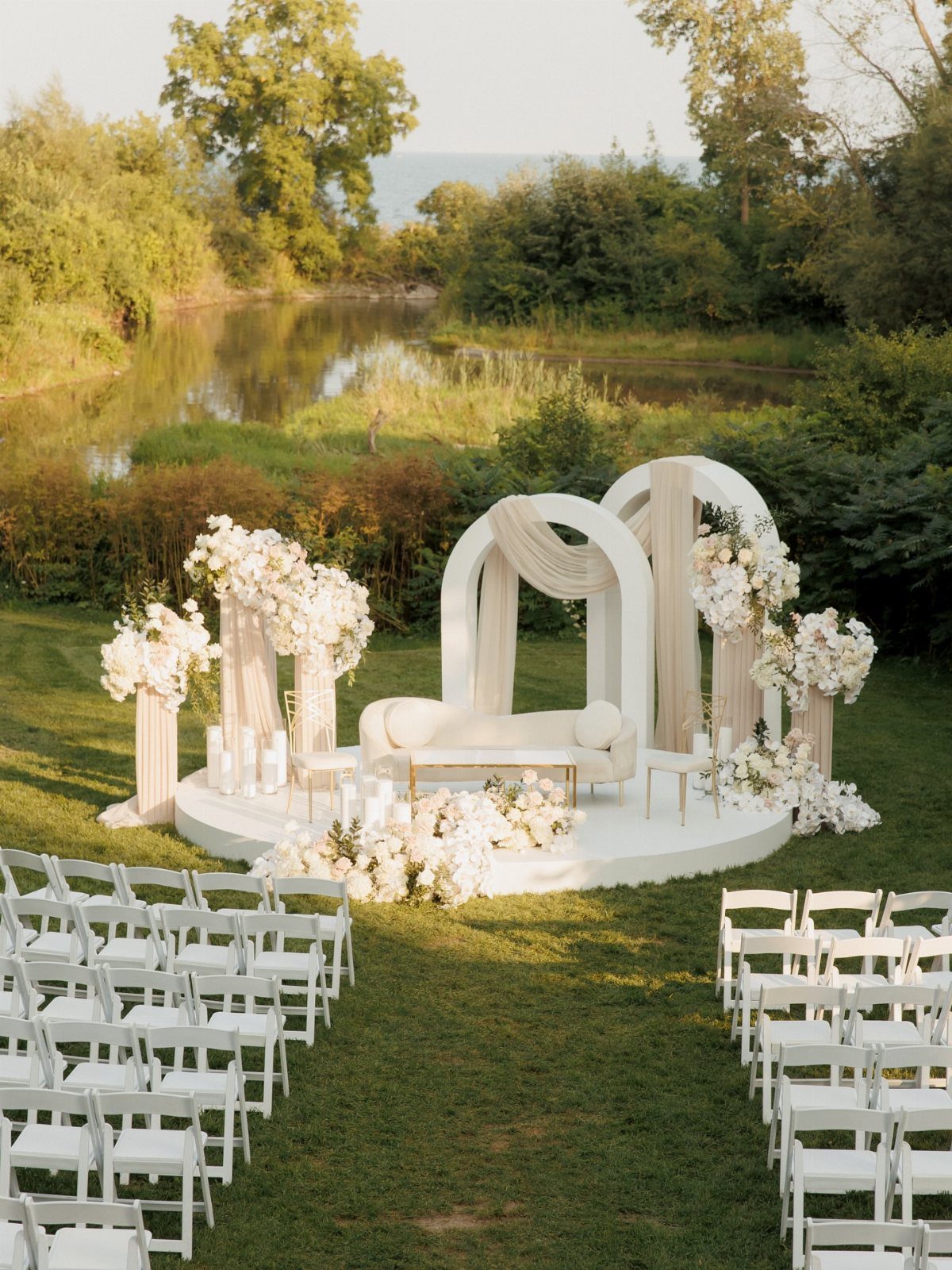 White wedding ceremony setup with floral arches and seating beside a peaceful river.