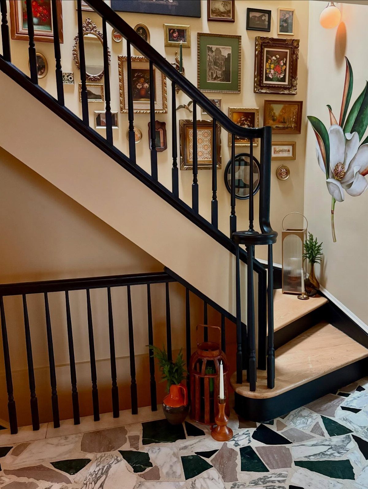 Framed art and mirrors decorating a cream wall along a black stairwell railing.