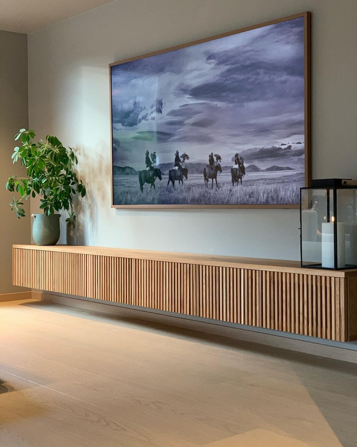 39 TV Design Ideas, from Built-In Shelving to Fireplace Walls 26 Light wood floating TV unit with vertical slats under a large framed landscape painting.