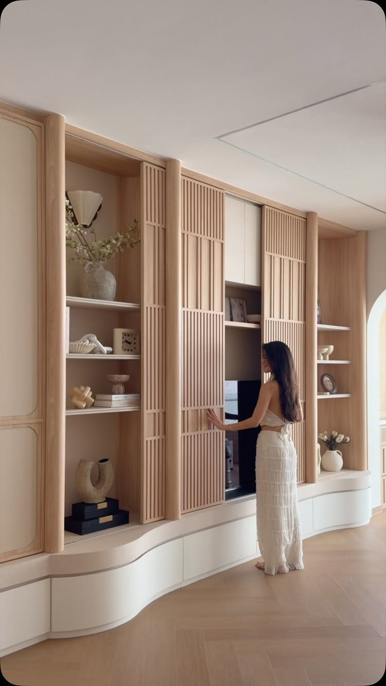 39 TV Design Ideas, from Built-In Shelving to Fireplace Walls 30 Woman standing by a light wood built-in TV unit with sliding slatted panels.