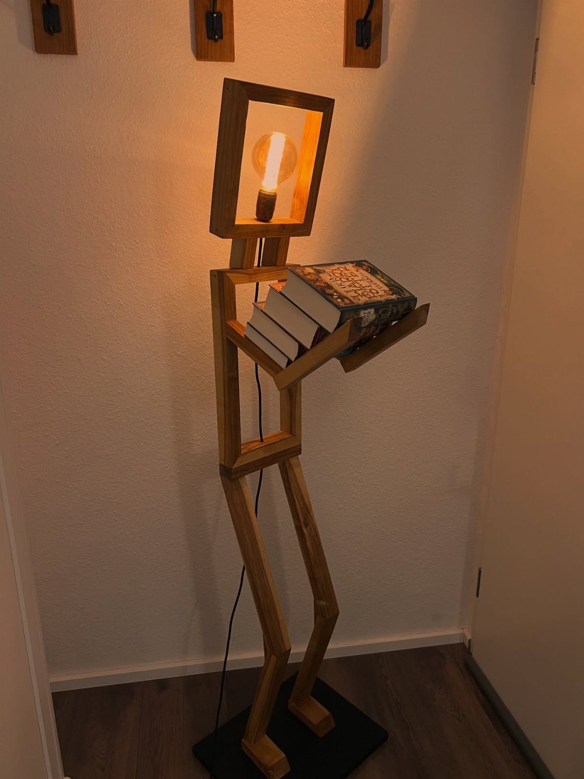 Humanoid wooden lamp with a glowing bulb head holding a shelf of books.