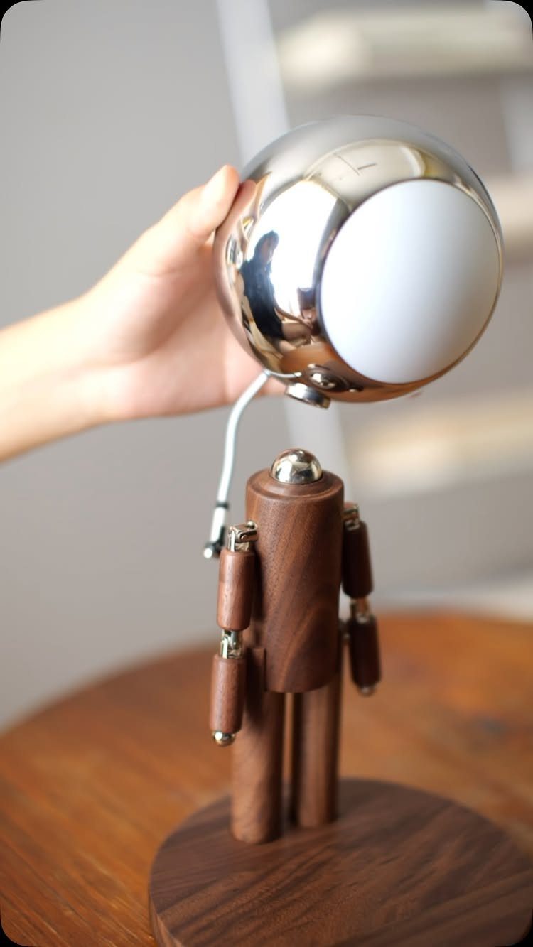 Wooden robot lamp with a chrome spherical head being held by a hand.